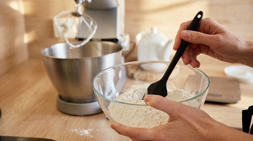Pessoa preparando ingredientes em tigela de vidro com espátula e batedeira planetária na bancada da cozinha